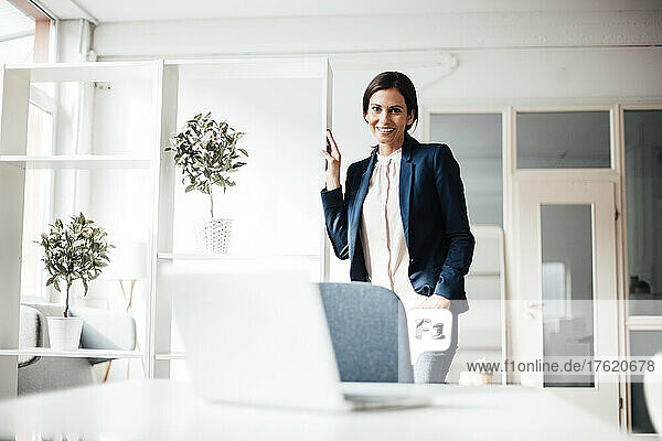 Happy businesswoman standing with hand in pocket at office