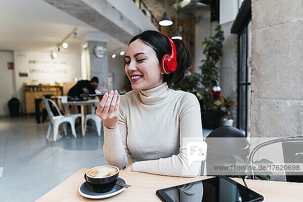 Smiling woman wearing headphones sending voicemail through smart phone at cafe