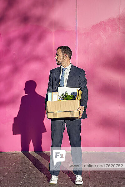 Businessman carrying box with personal belongings in front of pink wall