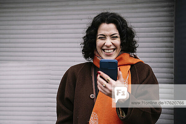 Happy young woman with mobile phone standing in front of corrugated shutter
