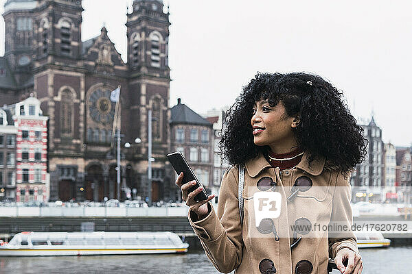 Smiling young woman with curly hair holding mobile phone in city