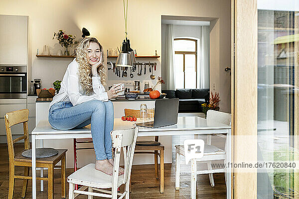 Glückliche blonde Frau hält eine Kaffeetasse und sitzt mit dem Kopf in der Hand auf dem Tisch