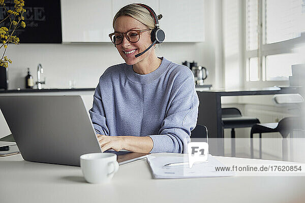 Smiling businesswoman wearing headset using laptop sitting at table