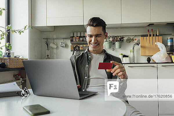 Happy young man with credit card doing online shopping through laptop in kitchen at home
