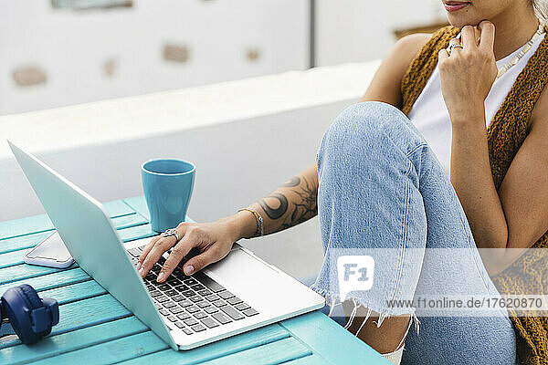 Young freelancer with tattoo typing on laptop sitting at table on rooftop
