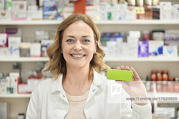 Lächelnde Apothekerin hält grüne Schachtel mit Medikamenten in der Apotheke