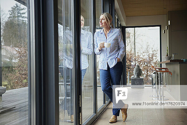 Frau hält Kaffeetasse und schaut mit der Hand in der Tasche durch das Fenster