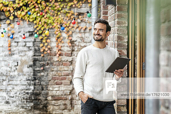 Smiling young holding tablet PC standing with hand in pocket by brick wall