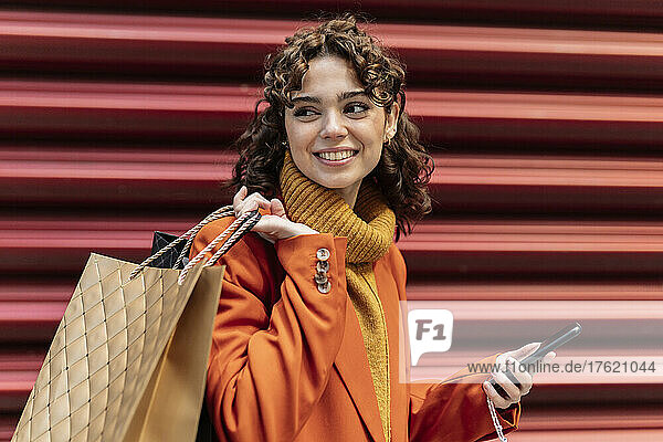 Young woman with smart phone and shopping bags by corrugated wall