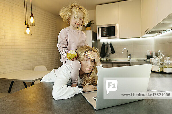 Tired woman with daughter looking at laptop in kitchen at home