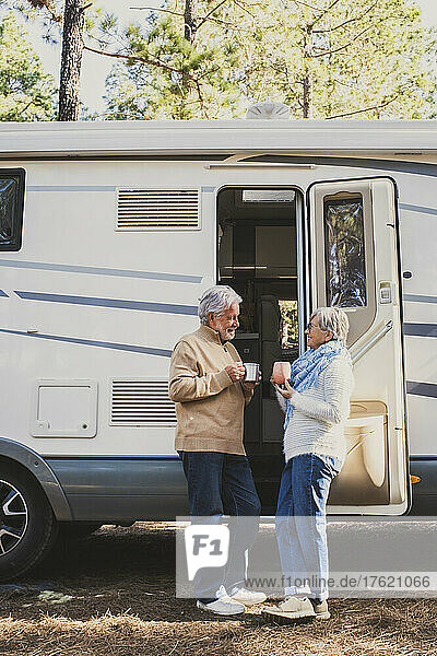 Senior couple with coffee cup standing by motor home in forest
