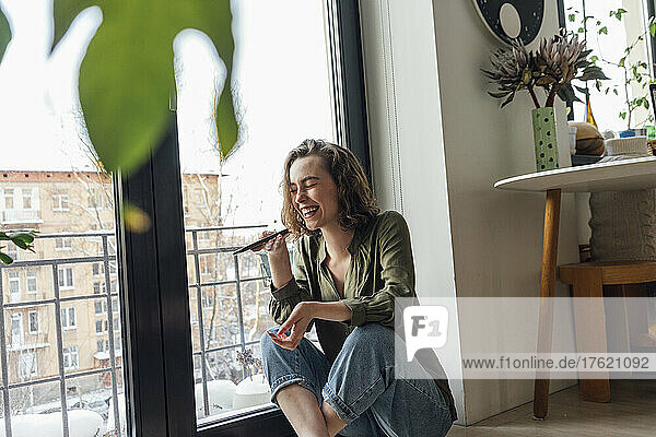 Cheerful woman with eyes closed sending voicemail through smart phone by glass window at home