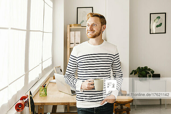 Smiling young man holding coffee cup standing with hand in pocket at home