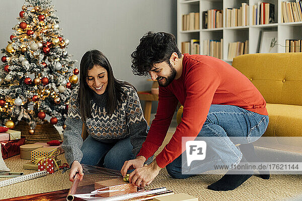 Smiling couple wrapping Christmas present in living room at home