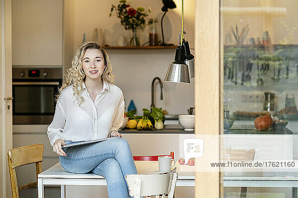 Young blond woman with laptop sitting on table in kitchen