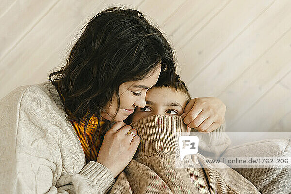 Smiling mother covering boy face with sweater at home