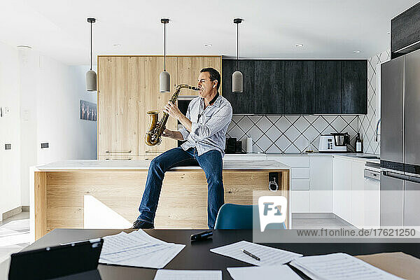 Musician with eyes closed playing saxophone sitting on kitchen island at home