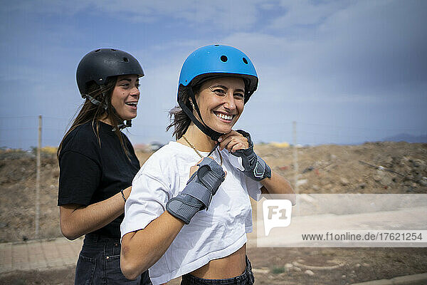 Happy young woman adjusting helmet by friend on sunny day