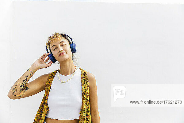 Smiling woman with eyes closed listening music on headphones in front of white wall