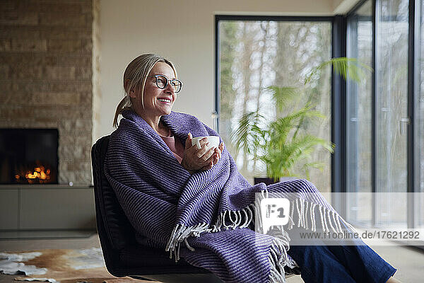Happy blond woman wrapped in blanket holding coffee cup sitting at home