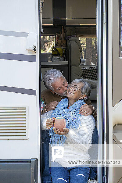 Senior man kissing happy woman on cheek sitting at door of motor home