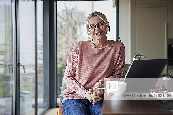 Smiling blond woman sitting with laptop in kitchen at home