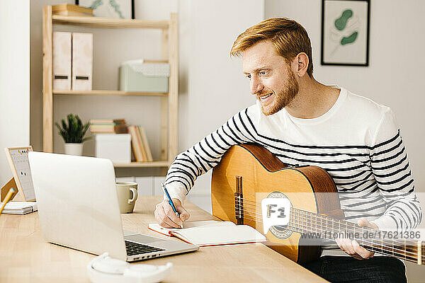 Lächelnder Mann mit Gitarre schreibt E-Learning-Musiknoten über Laptop und sitzt am Tisch