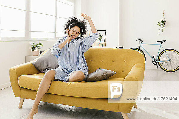 Happy young woman with hand raised enjoying music through wireless headphones sitting on sofa