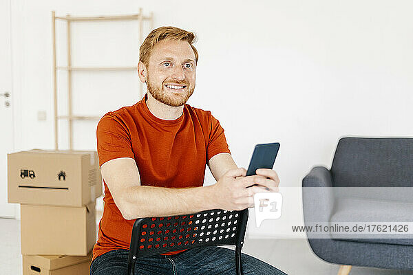Happy young man holding mobile phone sitting on chair at home
