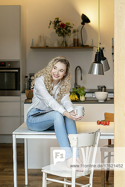 Lächelnder blonder Freiberufler mit Kaffeetasse und Laptop auf dem Tisch in der heimischen Küche