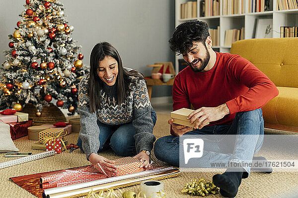 Happy couple wrapping Christmas present sitting in front of sofa
