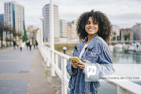Lächelnde Frau mit Smartphone lehnt am Geländer in der Stadt