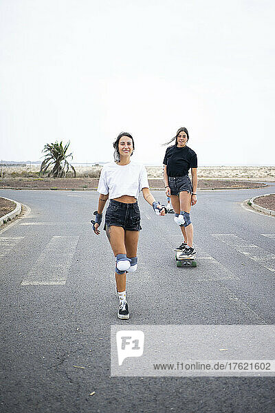 Young woman walking on road with friend skateboarding in background