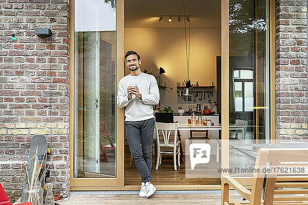 Smiling young man holding glass of coffee leaning on doorway