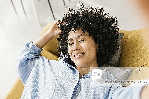 Smiling young woman with black curly hair lying on sofa at home