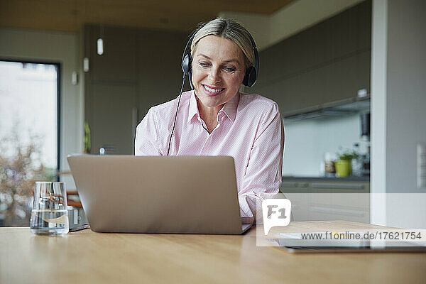 Smiling blond woman wearing headphones using laptop sitting at table