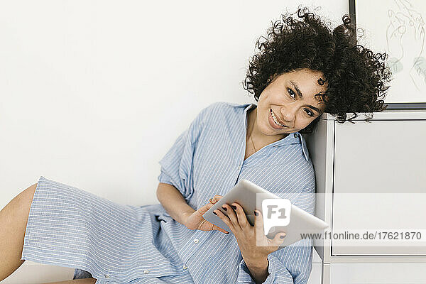 Happy young woman with tablet PC leaning on cabinet at home