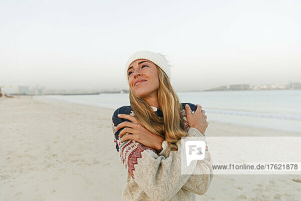 Smiling woman in warm clothing hugging self at beach