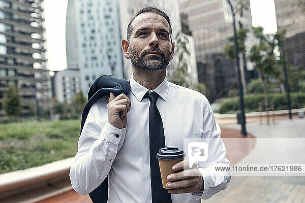 Portrait of businessman standing outdoors with jacket and disposable coffee cup in hands