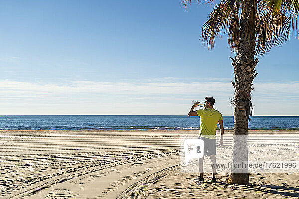 Man drinking water standing by palm tree at beach on sunny day