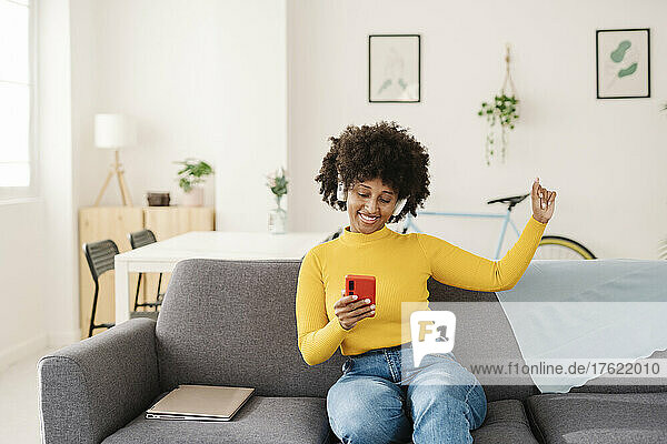 Smiling young woman using smart phone enjoying music through wireless headphones on sofa at home