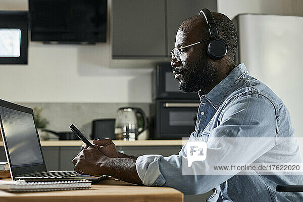Freelancer with headphones using smart phone by laptop sitting at home