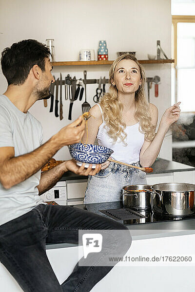 Young man looking at girlfriend tasting food in kitchen