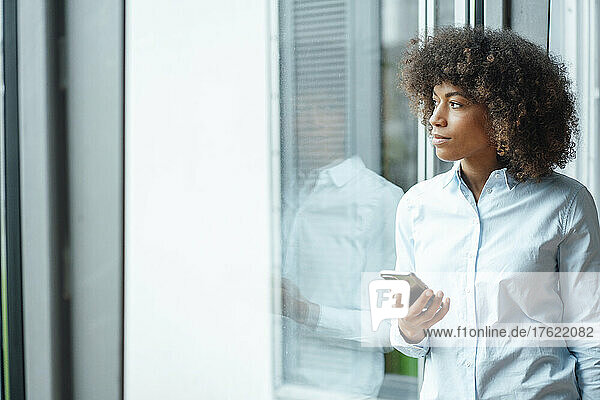 Businesswoman with mobile phone looking through window in office