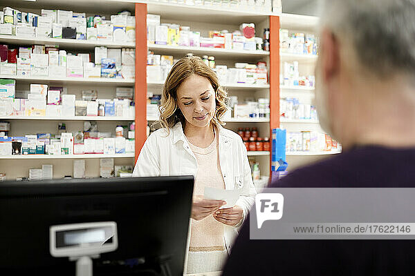 Female pharmacist reading prescription to customer at checkout counter in pharmacy store