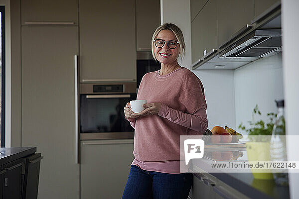 Happy blond woman holding coffee cup standing by kitchen counter at home