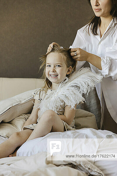 Woman tying hair of cheerful daughter sitting with feather on bed at home
