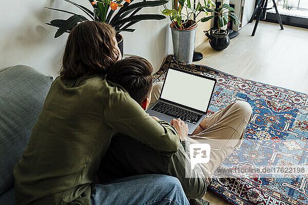Young woman sitting with arm around boyfriend using laptop at home