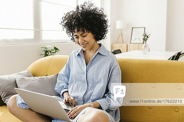 Lächelnde junge Frau sitzt mit Laptop auf dem Sofa im Wohnzimmer