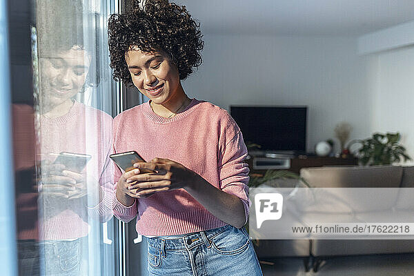 Lächelnde junge Frau  die ihr Mobiltelefon benutzt und sich zu Hause an ein Glasfenster lehnt